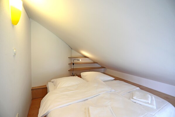 Un dormitorio con techo inclinado, una cama con ropa de cama blanca, una mesita de noche y una lámpara fijada a la pared.