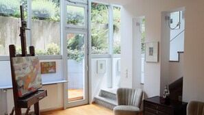 Una habitación con piso de madera, un cuadro sobre caballete, una silla, un aparador y un ventanal con vistas a la vegetación.