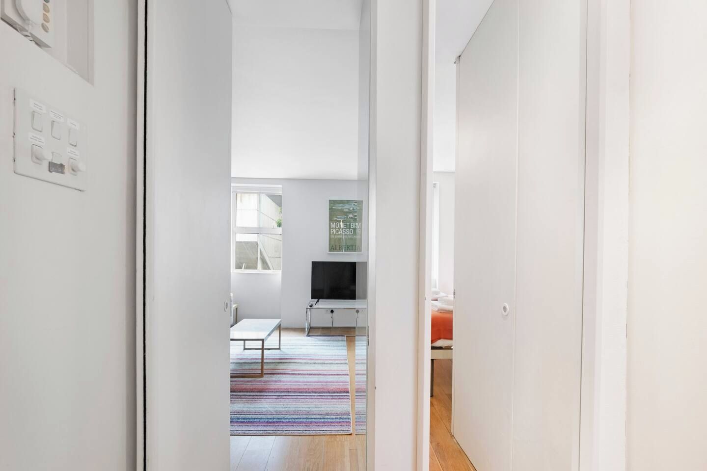 A modern interior with a hallway leading to a room with a television, a colorful rug, and a framed artwork on the wall.