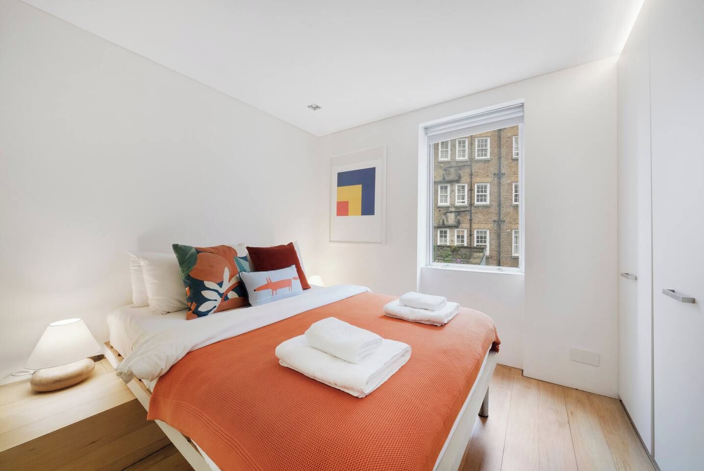Een moderne slaapkamer met een groot bed, een nachtkastje, een lamp en een raam met uitzicht op gebouwen.