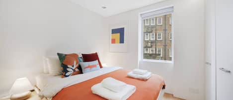 A modern bedroom with a large bed, a nightstand, a lamp, and a window with a view of buildings.
