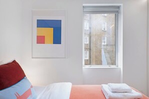Un dormitorio con cama, almohadas, una obra de arte enmarcada y una ventana con vista a edificios.