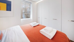 A modern hotel room with a large bed, white wardrobe, and a window with a view of buildings.