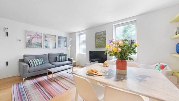 Ein modernes Wohnzimmer mit einem grauen Sofa, einem Streifen Teppich, einem Esstisch mit Stühlen, einer Blumenvase und Kunstwerken an den Wänden.