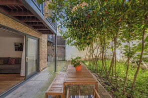 Un patio moderno al aire libre con una mesa y banco de madera, rodeado de exuberante follaje y un muro de ladrillo.