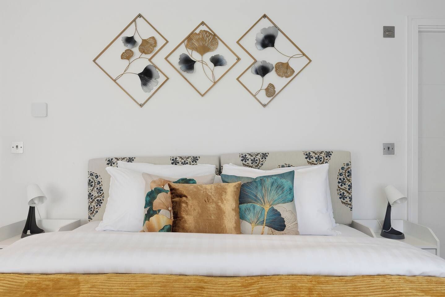 Un llit amb capçalera de fusta, roba de llit blanca i almoiners decoratius amb motius florals.