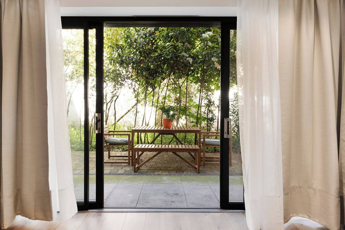 Una habitación con puerta corrediza de vidrio que da a un patio exterior con mesa y sillas de madera, una planta en maceta y suelo de ladrillo.