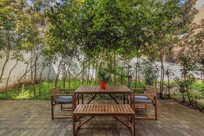 An outdoor patio with a wooden table and benches, surrounded by lush greenery and trees.