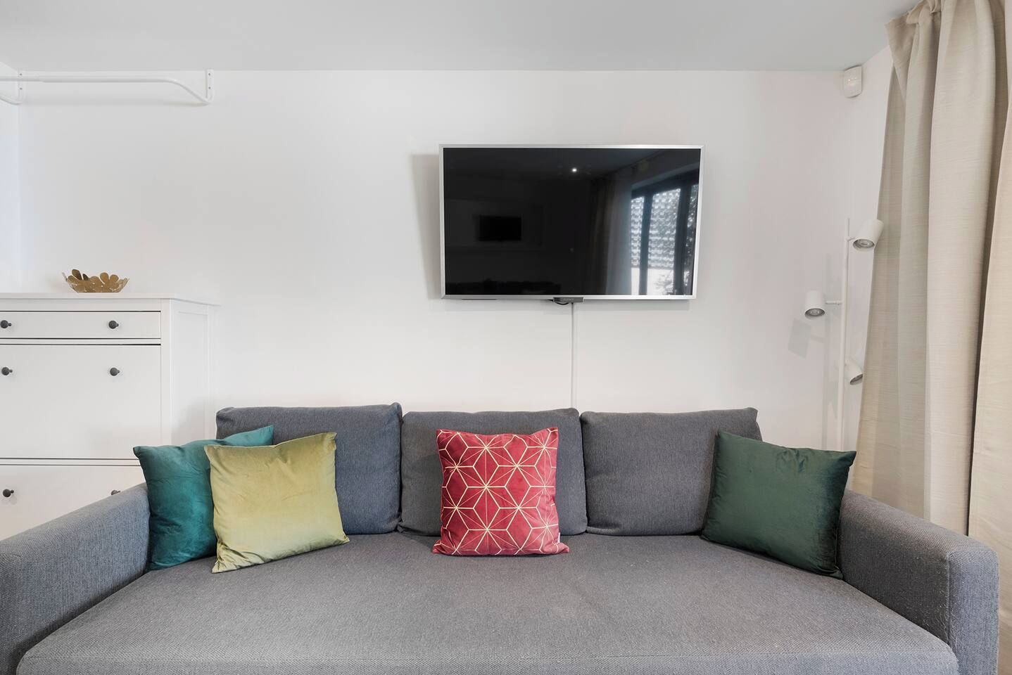 A living room with a grey sofa, a flat-screen TV mounted on the wall, and a white dresser with drawers.