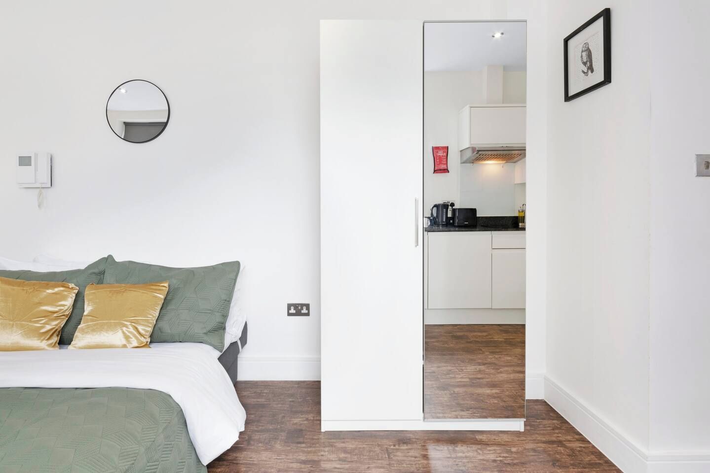 A bedroom with a bed, a green and gold pillow, a round mirror, and a white door leading to a kitchen.