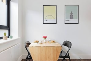 Uma área de refeições com uma mesa de madeira, duas cadeiras, uma janela com um cacto e duas fotografias emolduradas na parede.