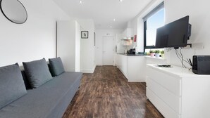 Ein modernes, helles Zimmer mit einer grauen Couch, einem Flachbildfernseher und einer Küchenzeile mit weißen Schränken und einem Fenster.