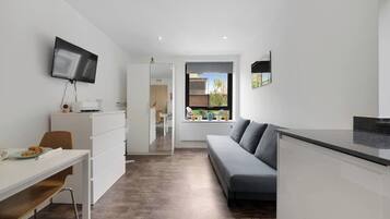 A modern, well-lit room with a flat-screen TV, a sofa, a dining table, and a view of the outdoors through a large window.