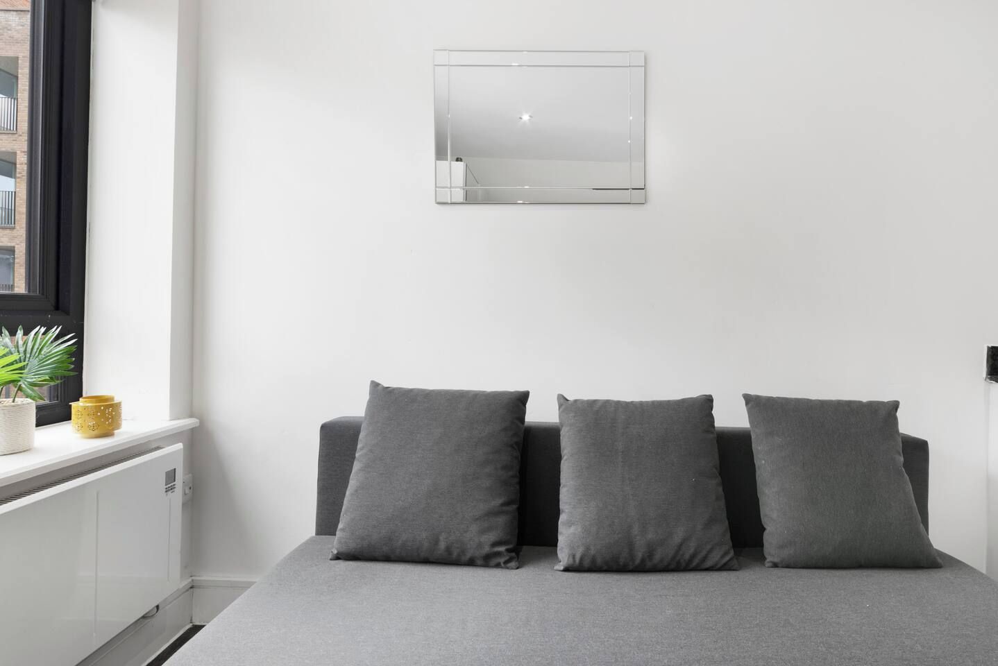 A minimalist bedroom with a bed, two pillows, a small window, and a wall-mounted mirror.