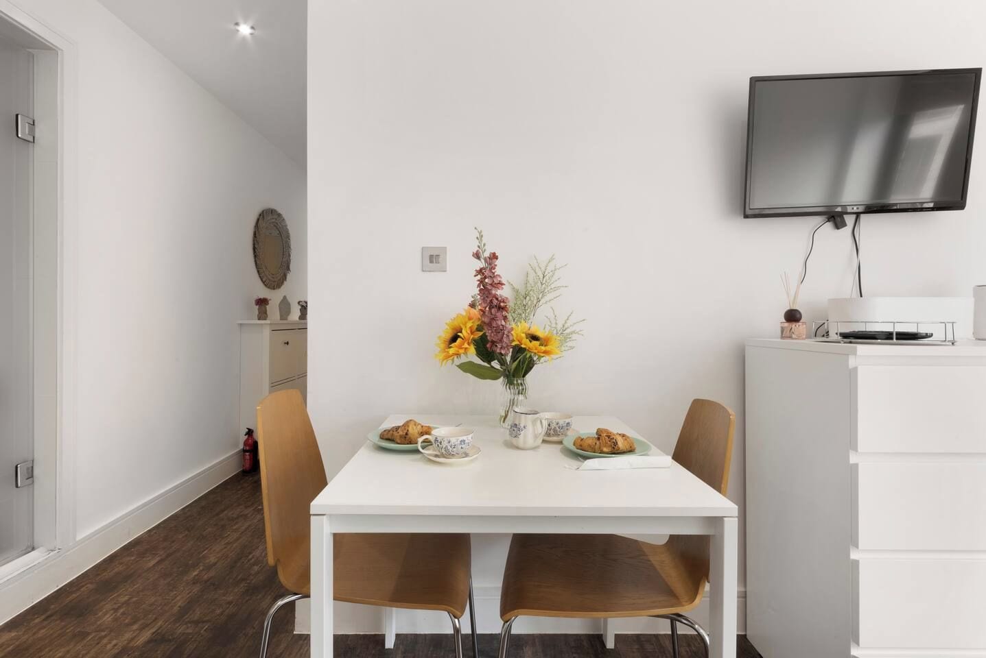 A modern dining area with a white table, two chairs, a vase of flowers, and a flat-screen TV mounted on the wall.