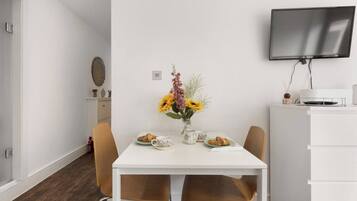 A modern dining area with a white table, two chairs, a vase of flowers, and a flat-screen TV mounted on the wall.