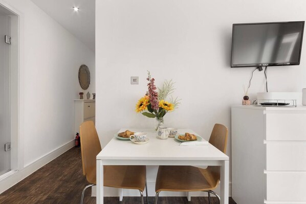 Un comedor moderno con una mesa blanca, dos sillas, un jarrón de flores y un televisor de pantalla plana montado en la pared.