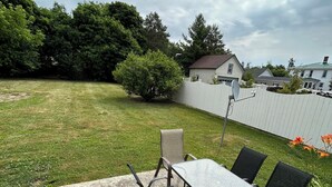 A patio with a table and chairs, a white fence, and a grassy yard with trees and a house in the background.