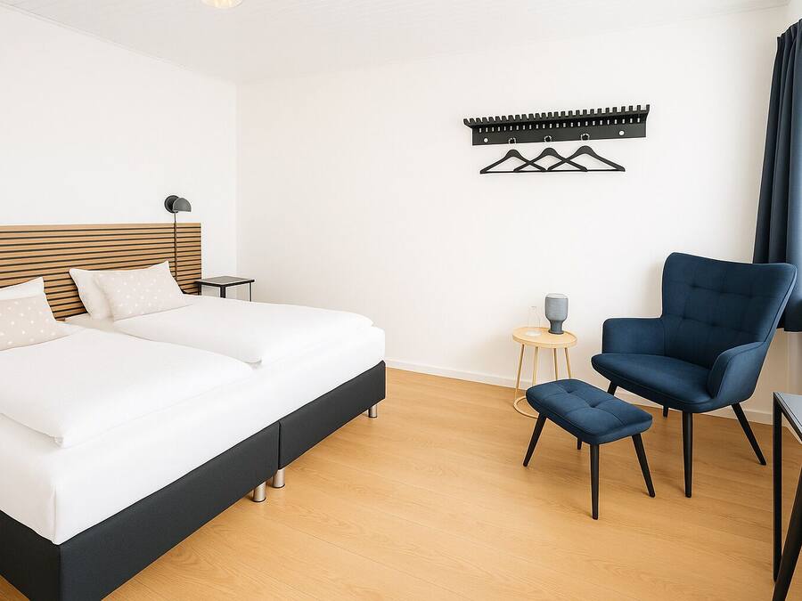 A modern bedroom with a large bed, a blue armchair, a small side table, and a wall-mounted coat rack.