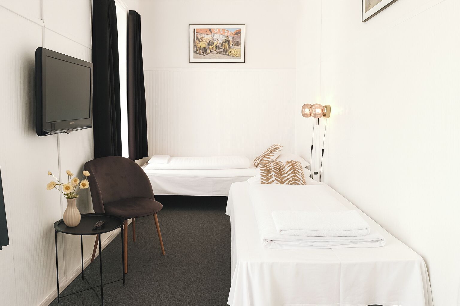 A hotel room with a bed, a chair, a small table with a vase, a flat-screen TV mounted on the wall, and a bedside lamp.