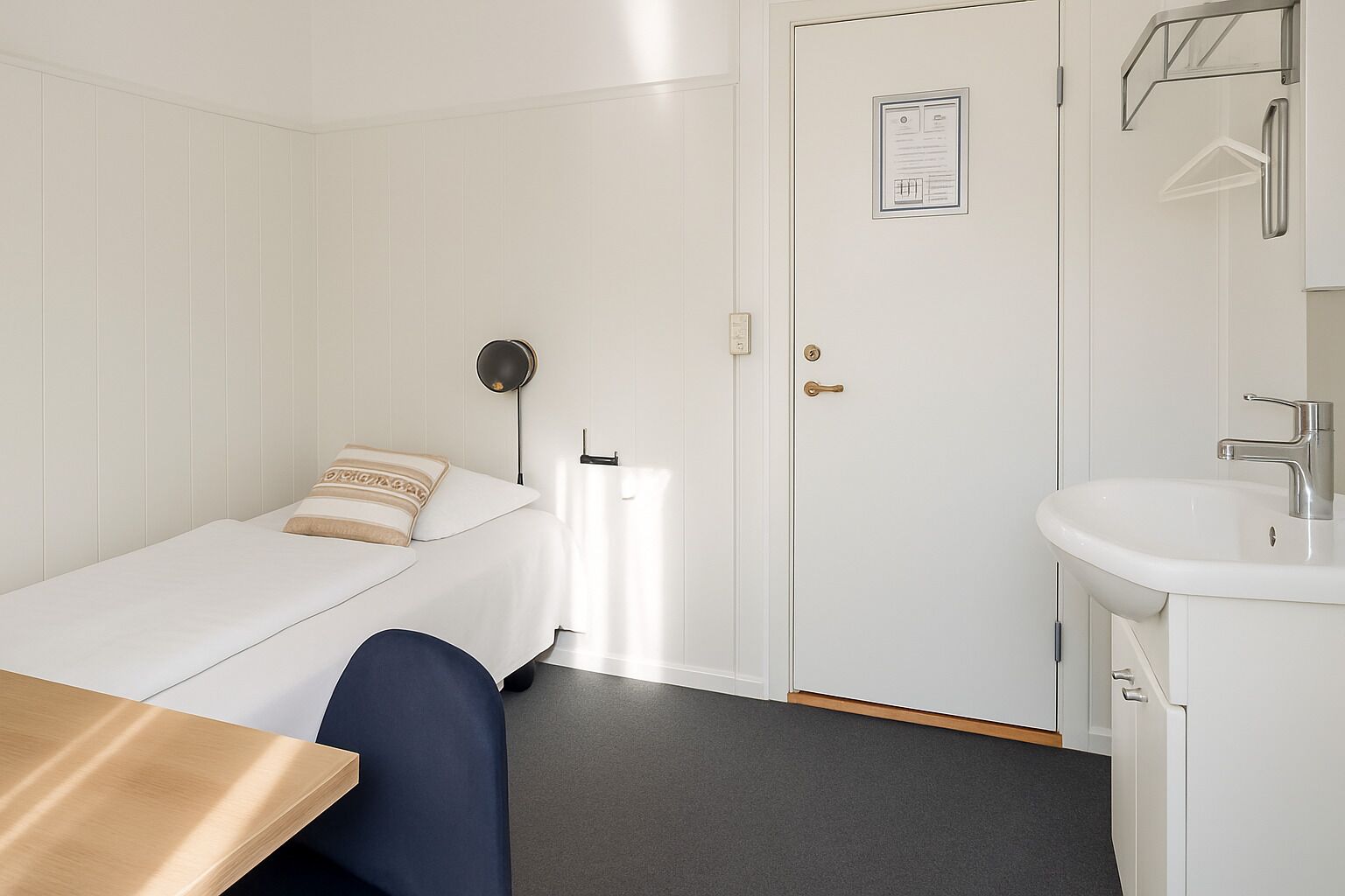 Una habitación pequeña y limpia con cama, escritorio, lavamanos blanco y una puerta con un certificado.