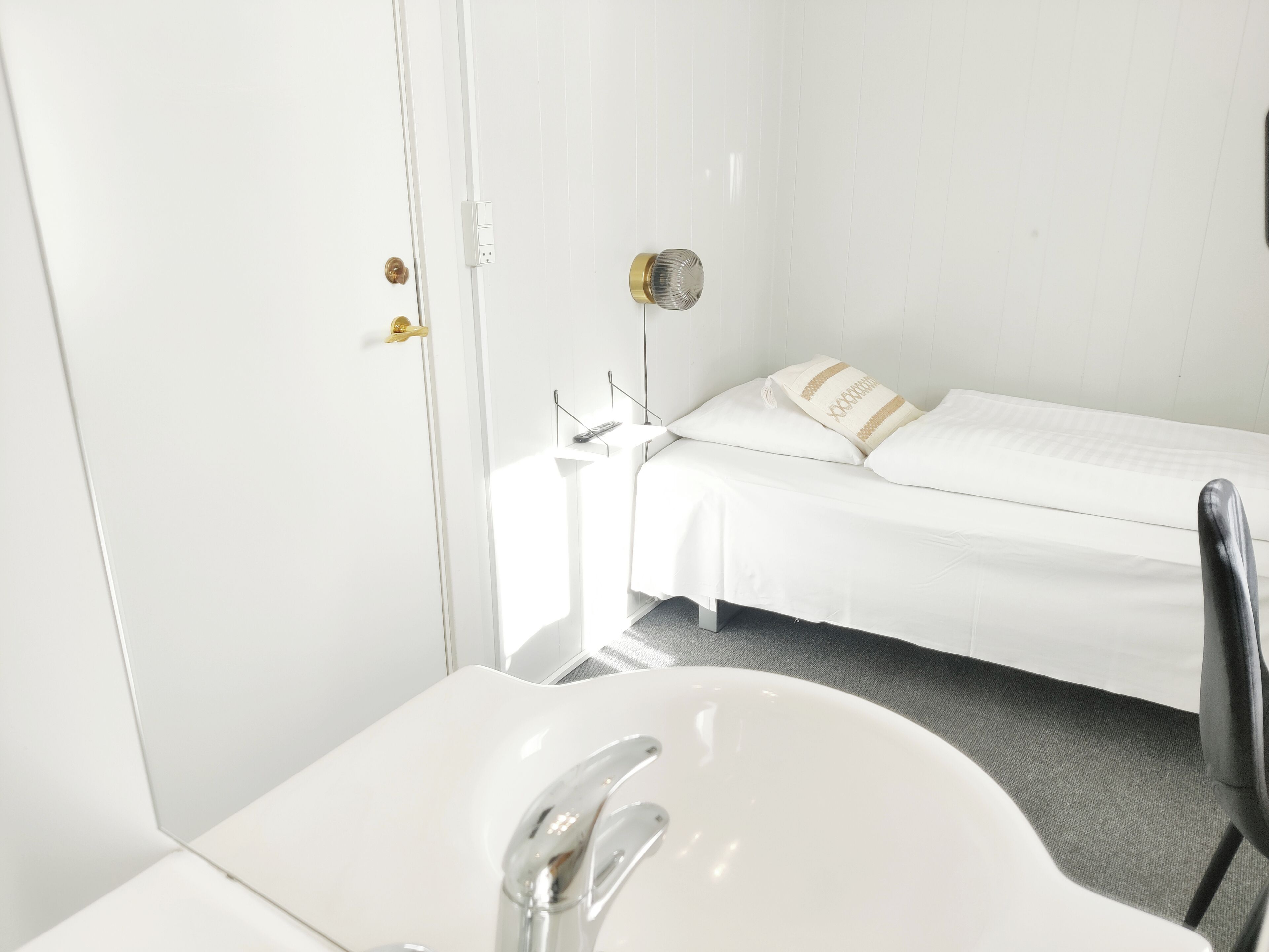Une petite pièce propre, avec un lit simple, un lavabo blanc et une porte dotée d’une poignée.