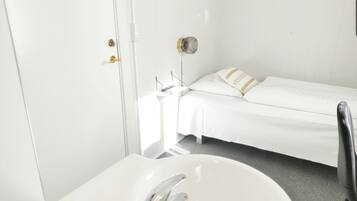 Une petite pièce propre, avec un lit simple, un lavabo blanc et une porte dotée d’une poignée.