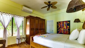 A bedroom with a bed, wooden wardrobe, ceiling fan, and artwork on the wall. - TBS 39 Beachside-Resort Style Condo-Pool-1 Bedroom (Placencia)
