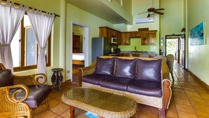 A spacious living room with a leather sofa, wicker chairs, and a glass coffee table. A kitchen is visible in the background with wooden cabinets and a microwave. - TBS 39 Beachside-Resort Style Condo-Pool-1 Bedroom (Placencia)