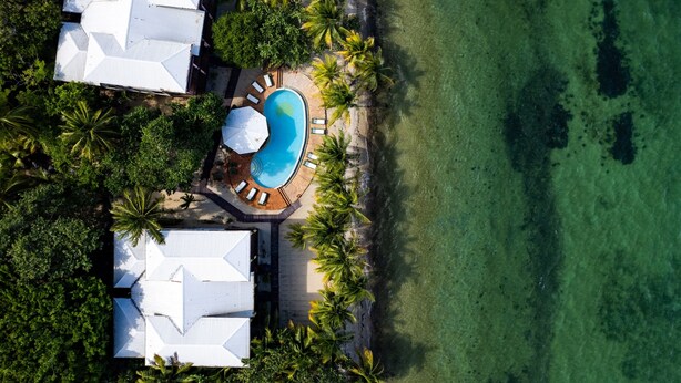 Eine Luftaufnahme eines Resorts mit einem Swimmingpool, umgeben von Palmen und Liegestühlen, direkt neben einem Sandstrand.