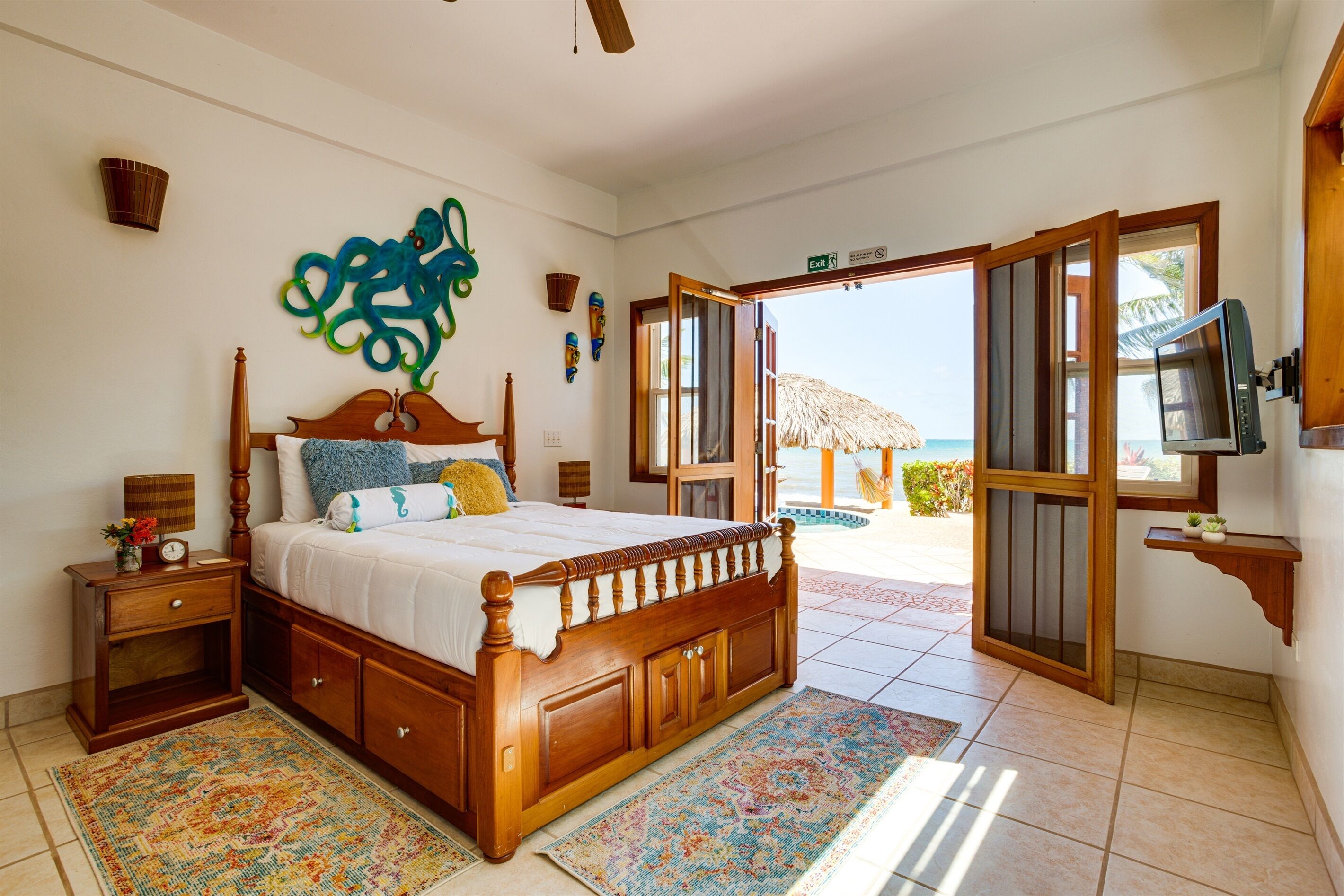 A bedroom with a wooden bed, a nightstand, a TV, and a view of the beach through an open door.