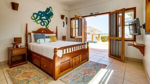 Un dormitorio con una cama de madera, una mesita de noche, un televisor montado en la pared y vista al mar a través de una puerta abierta.