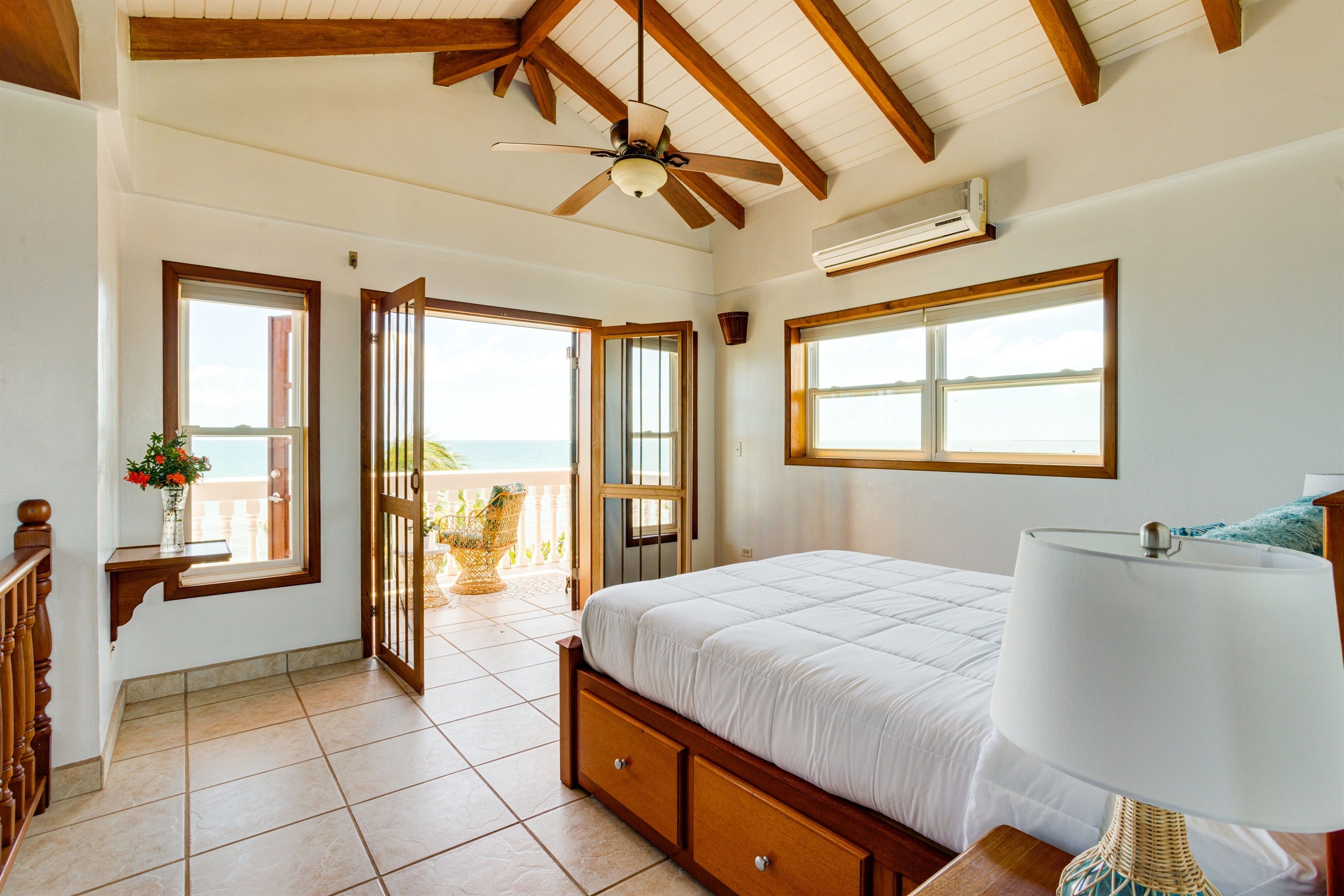 A bedroom with a bed, a ceiling fan, an air conditioning unit, and a view of the sea through open doors.