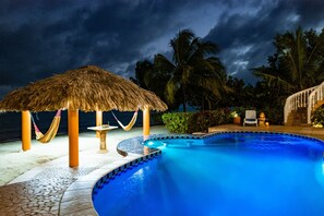 A swimming pool with a thatched gazebo, palm trees, and a view of the ocean. - Modern Beachfront Studio: Pool & Luxury (Placencia)
