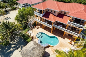 Ein großes, zweistöckiges Haus mit Swimmingpool, umgeben von Palmen und einem Reedschirm.