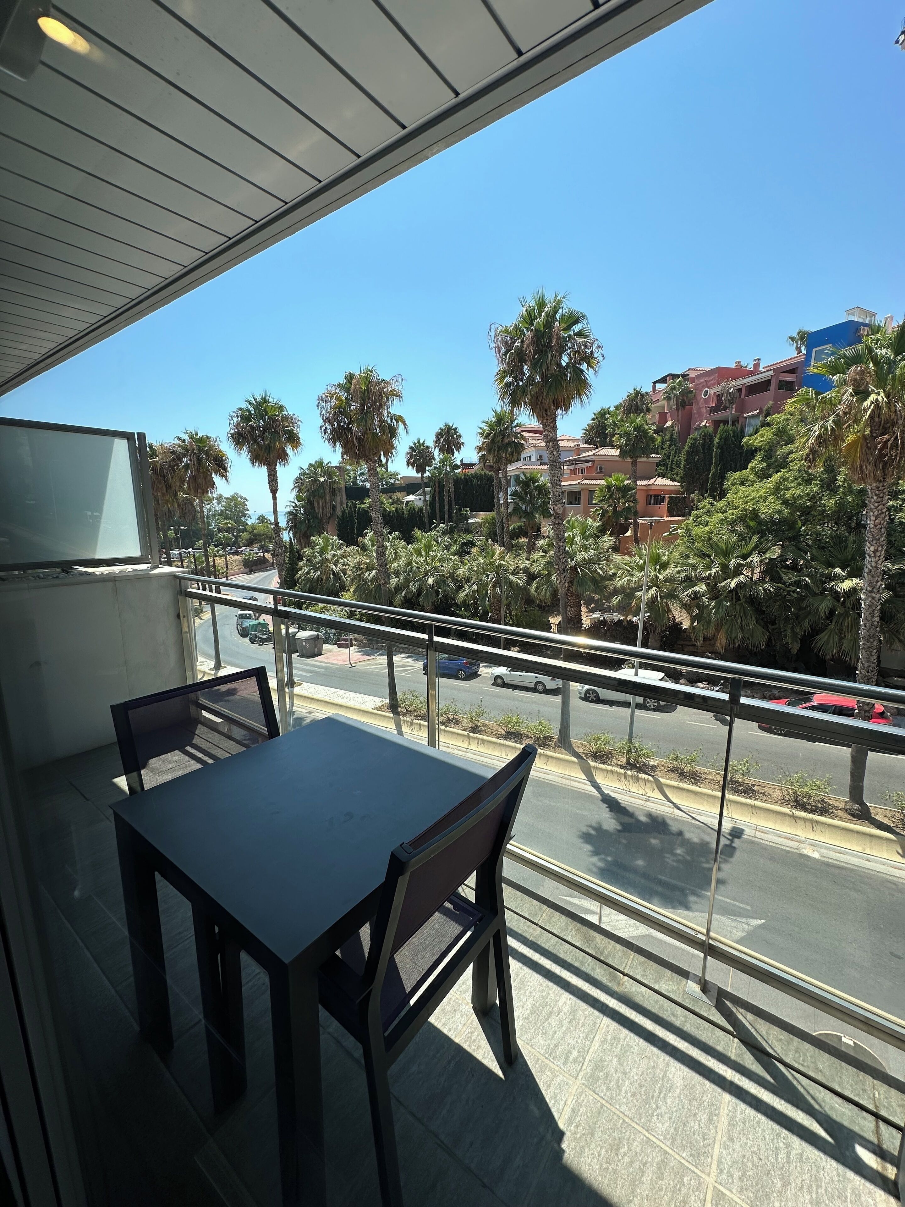 Een balkon met een tafel en stoelen, met uitzicht op een straat met palmbomen en gebouwen.