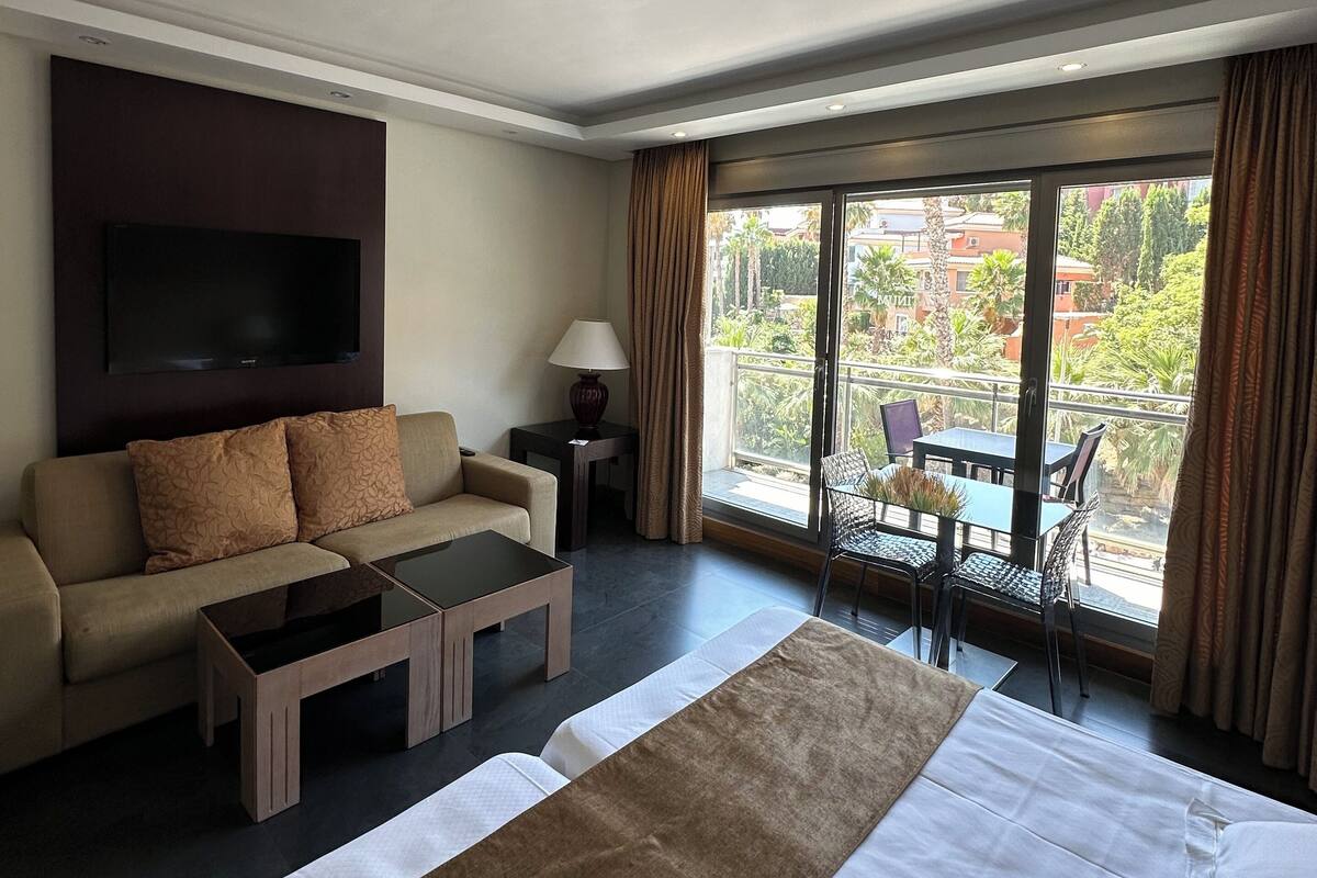 Ein Hotelzimmer mit Bett, Sofa, Couchtisch, Fernseher und einem Balkon mit Tisch und Stühlen.