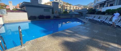 A swimming pool with a tiled deck, a small lounge area with sun loungers, and a view of surrounding buildings.