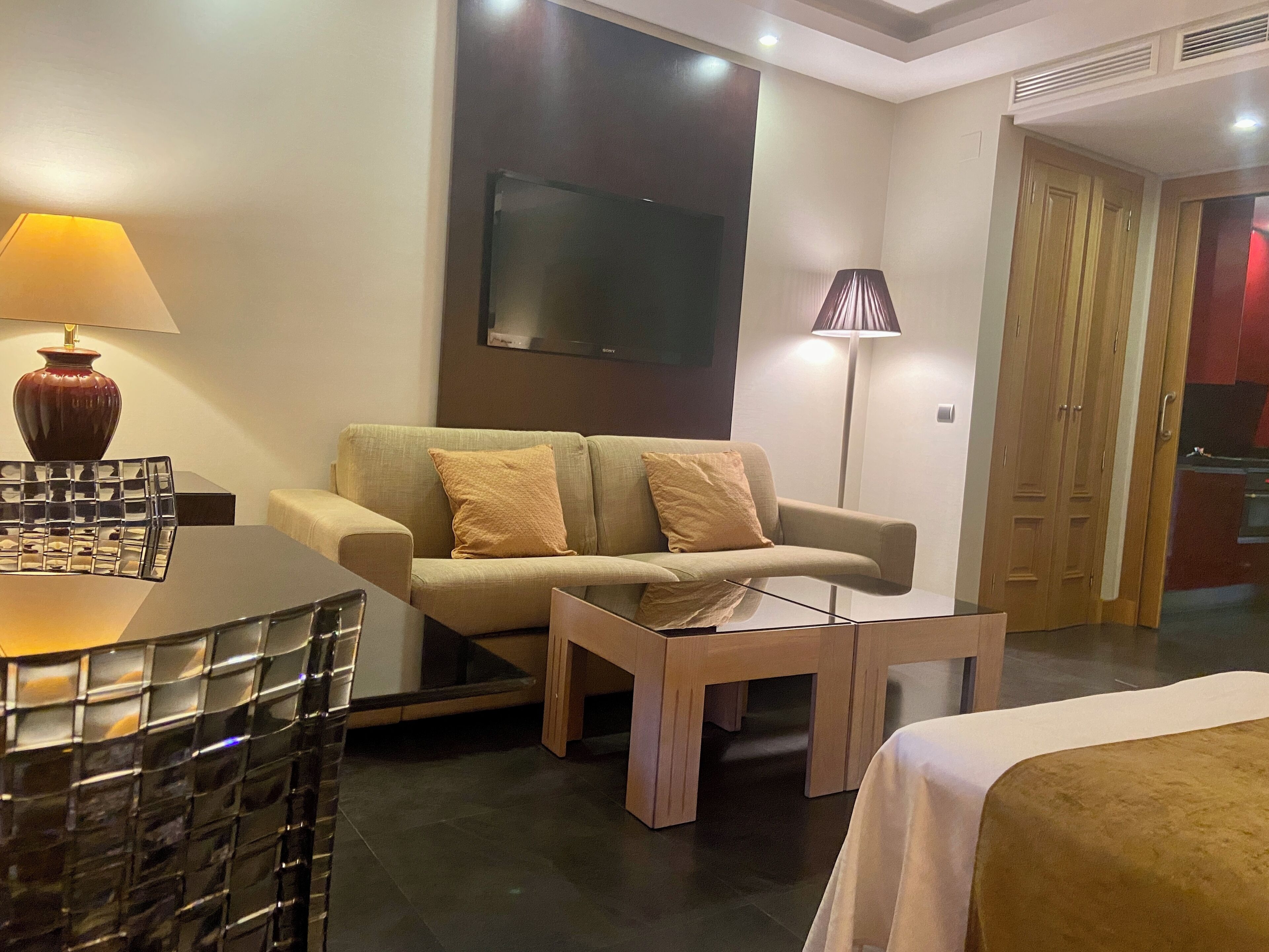 A modern hotel room with a sofa, a small coffee table, a TV mounted on the wall, and a wooden door.