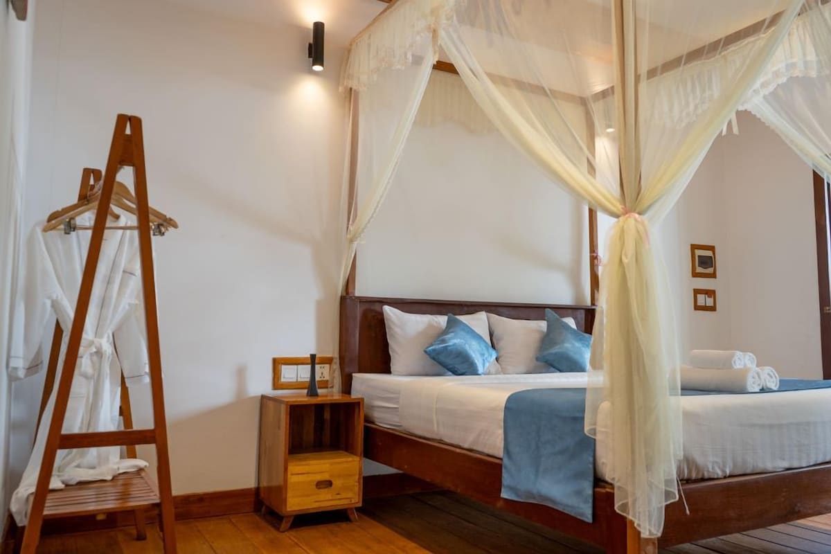 Ein Schlafzimmer mit einem hölzernen Bett, weißer Bettwäsche, einem Nachttisch aus Holz, einem Wäscheständer mit weißen Bademänteln und einem Baldachinbett.