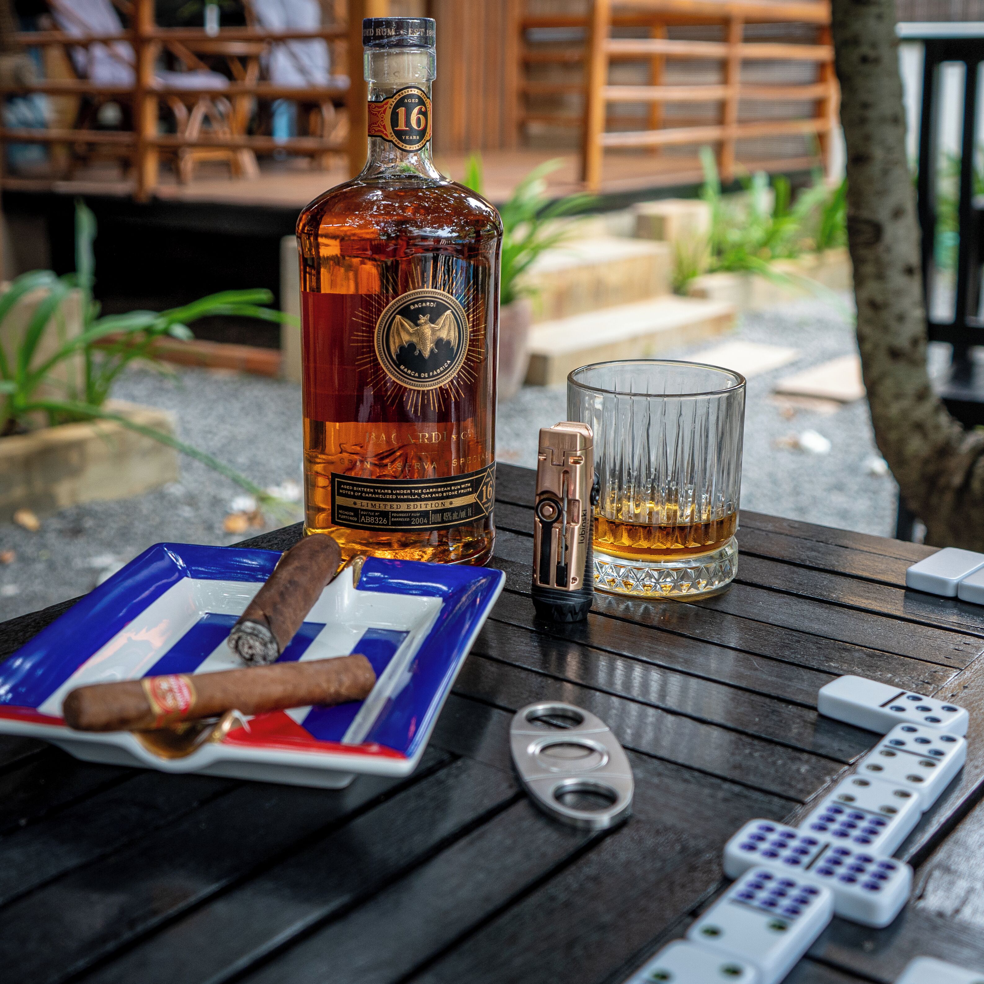 Una botella de ron añejo, un vaso, un encendedor, un cigarro, un control remoto y fichas de dominó sobre una mesa de madera.