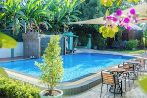 Une piscine dotée d’une terrasse en bois et de chaises, entourée d’une végétation luxuriante et d’un petit bâtiment.