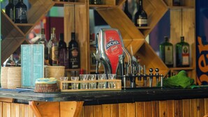 Uma área de bar com prateleiras de madeira repletas de garrafas de bebidas, um caixa registradora e utensílios de bar.