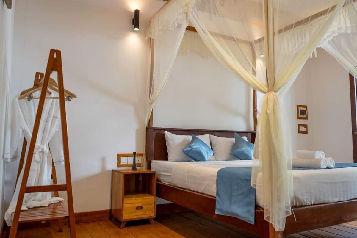 Un dormitorio con cama de madera, ropa de cama blanca, una mesita de noche de madera, un perchero de madera con batas blancas y un dosel sobre la cama.