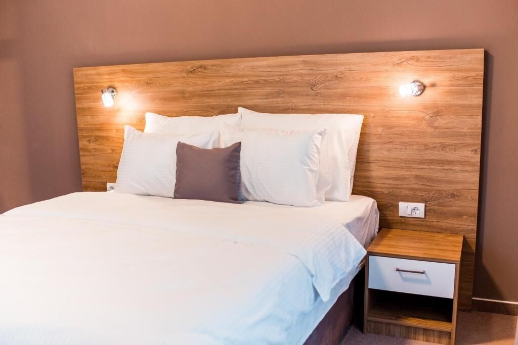 A neatly made bed with white linens and a wooden headboard. There is a small wooden nightstand with a drawer to the right of the bed.