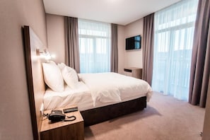 Une chambre d’hôtel avec un grand lit, une table de chevet avec un téléphone et une télécommande, un téléviseur fixé au mur et de grandes fenêtres avec des rideaux.