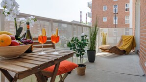 A wooden table with a bowl of fruit, a bottle, and two glasses of orange drink. There is a potted plant and a covered lounge chair. - Lumina deluxe 3-room apartment (Lodz)