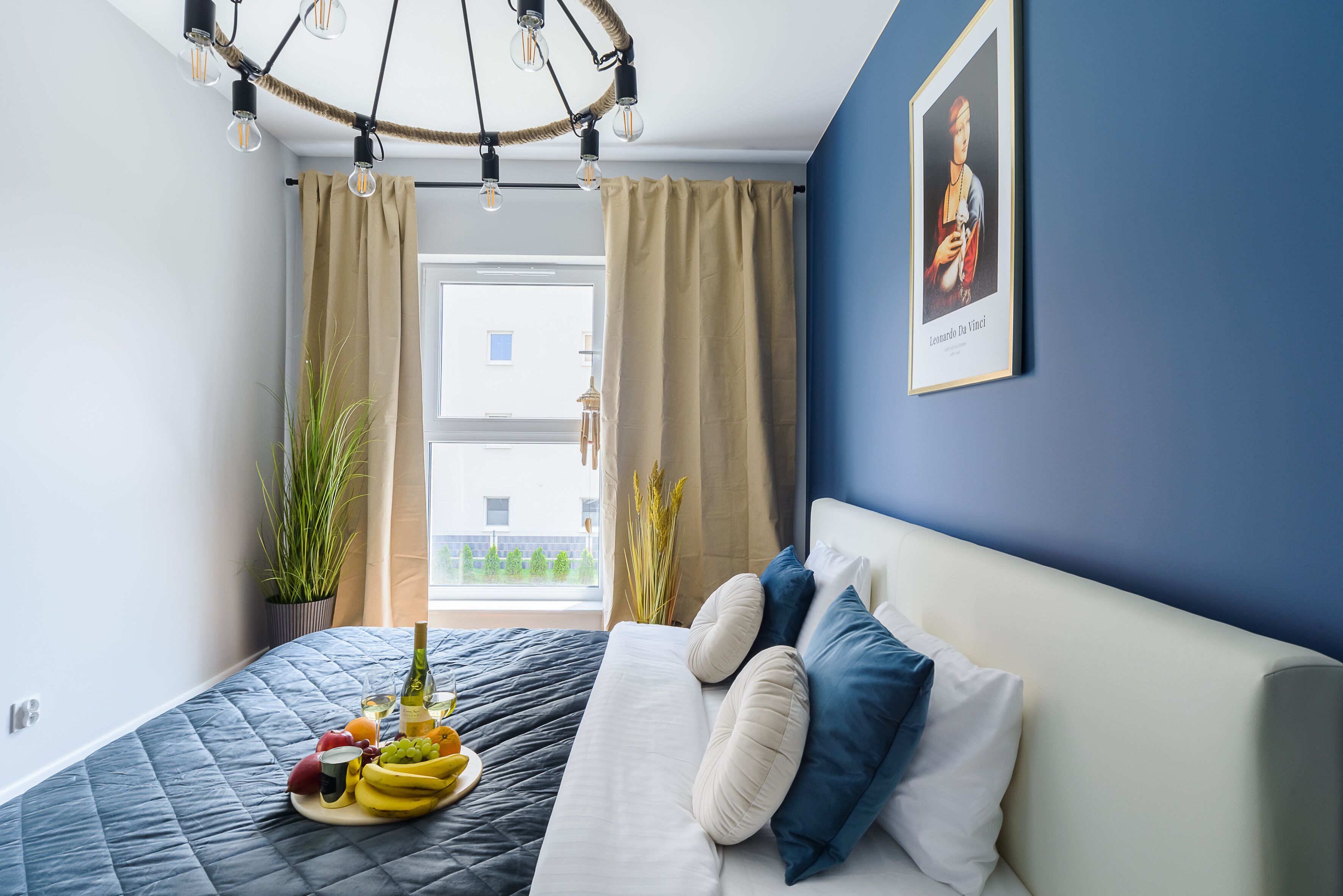 A bedroom with a bed, a bedside table with fruit, a hanging light fixture, and a window with curtains.