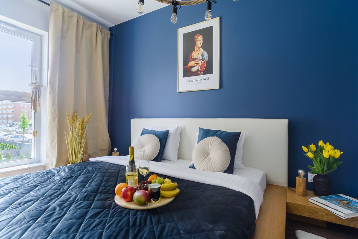 Un dormitorio con una cama, una mesita de noche con un libro, un florero con flores y una ventana con cortinas.