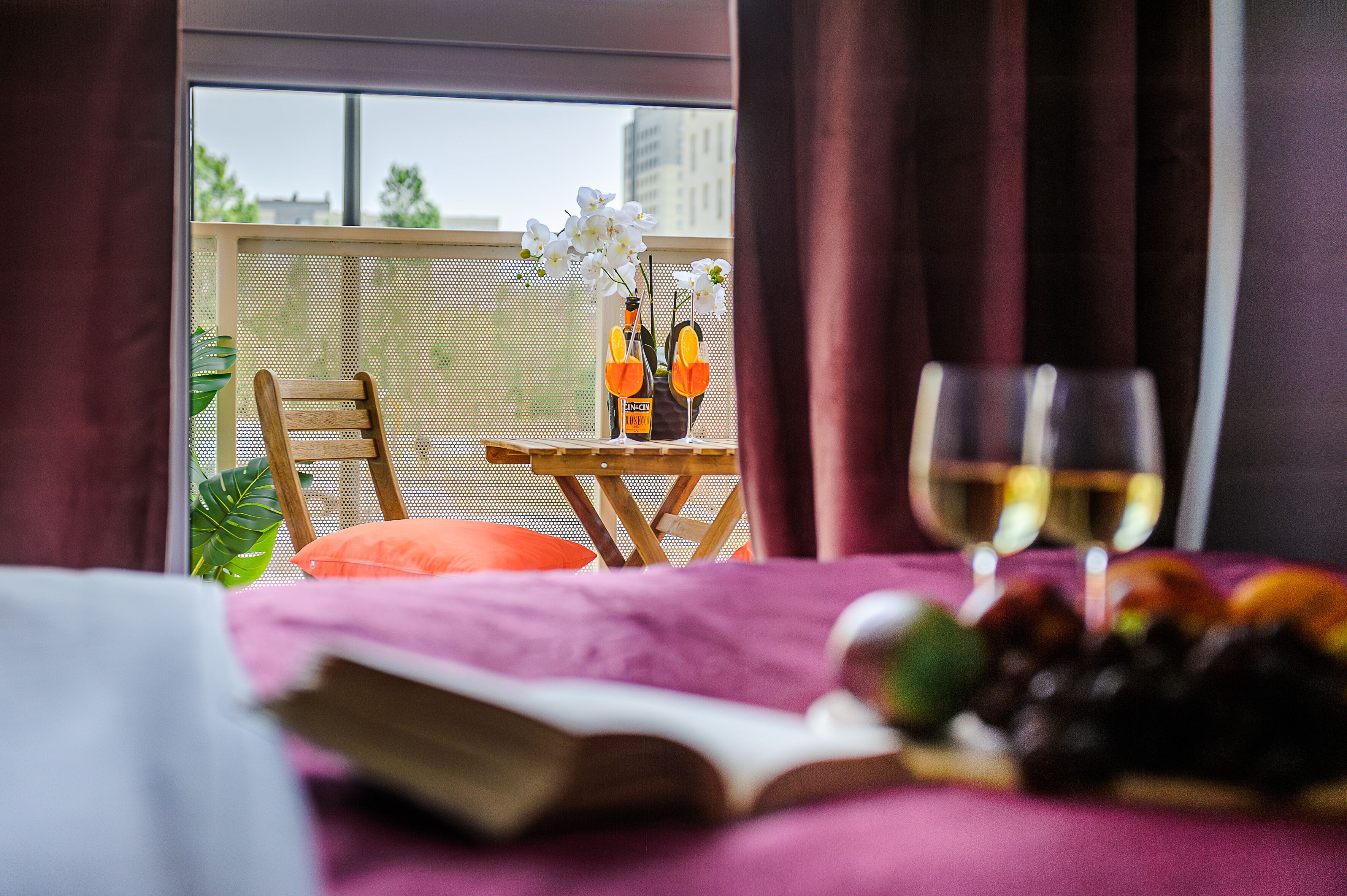 A room with a balcony, a table with a book, a glass of wine, and a fruit basket.